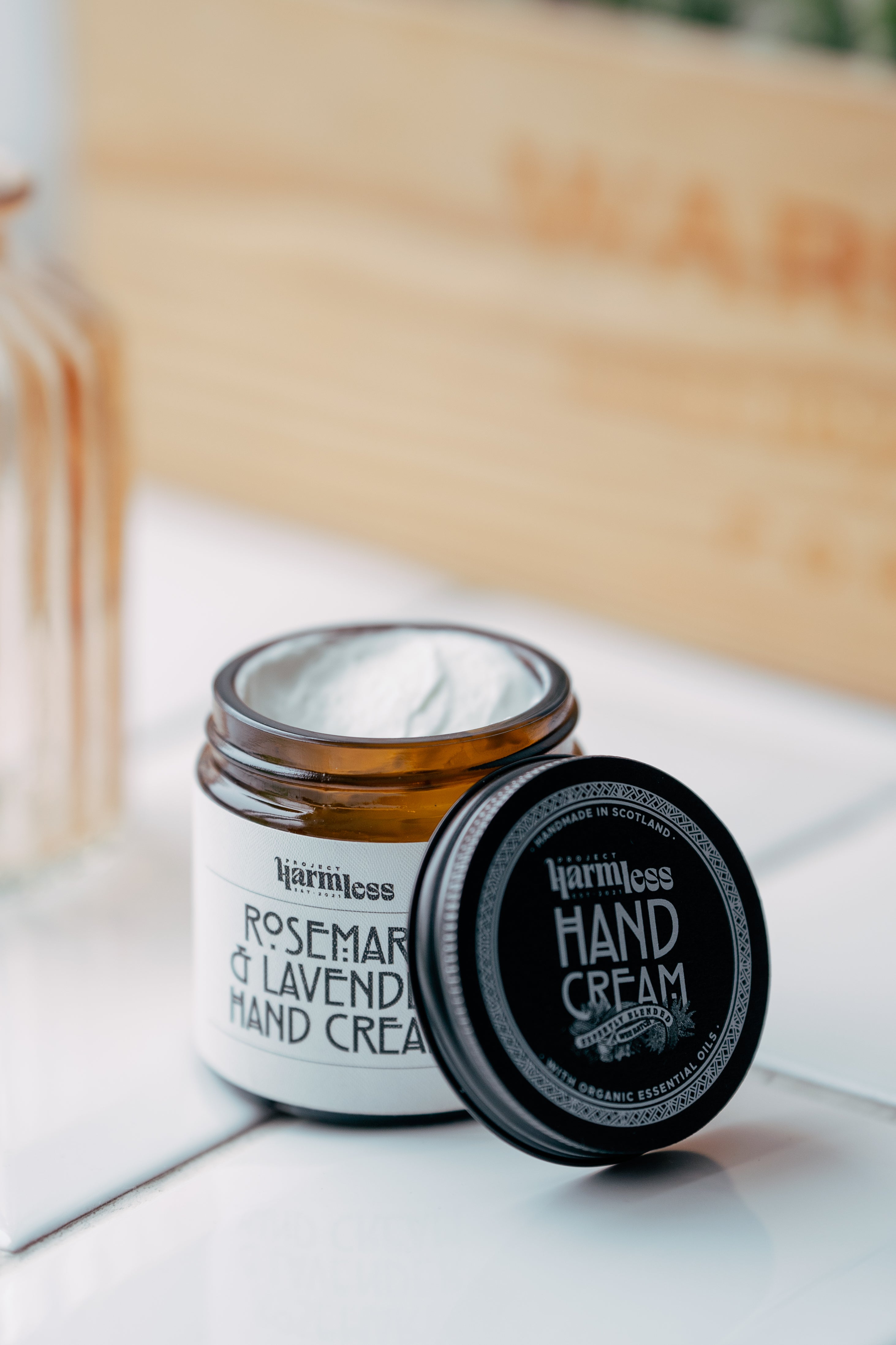 Project Harmless Rosemary and Lavender Hand Cream in a glass jar, placed on a kitchen countertop. The high-end, retro-designed black cap, featuring intricate Celtic knot details as a Scottish symbol, is positioned beside the jar. The cap includes the text ‘Handmade in Scotland’ and ‘With Organic Essential Oils,’ emphasising the brand’s artisanal quality. The luxurious design highlights ‘Expertly Blended’ and ‘Wee Batches’ for small-batch craftsmanship. A sophisticated, sustainable skincare choice.