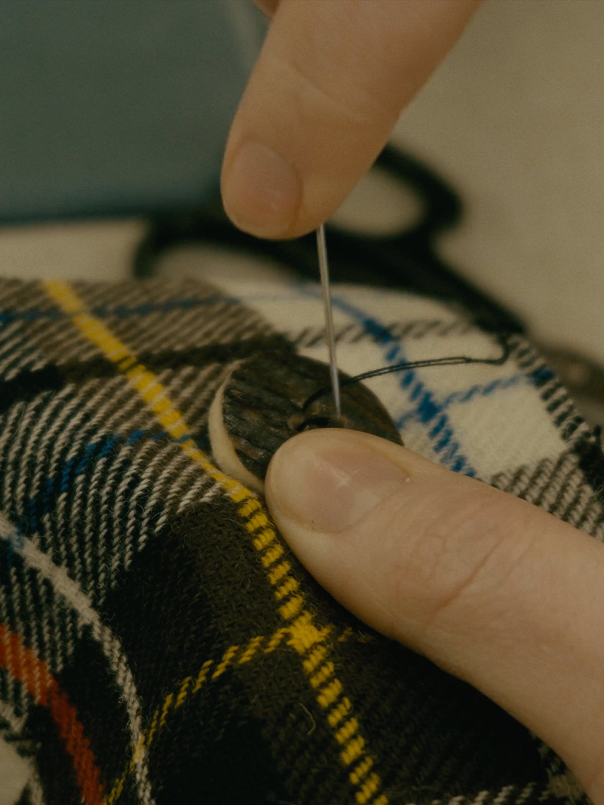 A close-up of hand-sewing a button onto the 1970 Commonwealth Games tartan at Harmless Atelier by Project Harmless in Scotland. Showcasing artisan craftsmanship, sustainable fashion, and plastic-free design.