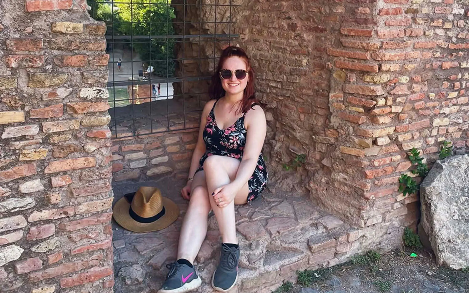 A woman sits in a stone archway surrounded by ancient brick walls, wearing sunglasses and a floral dress.