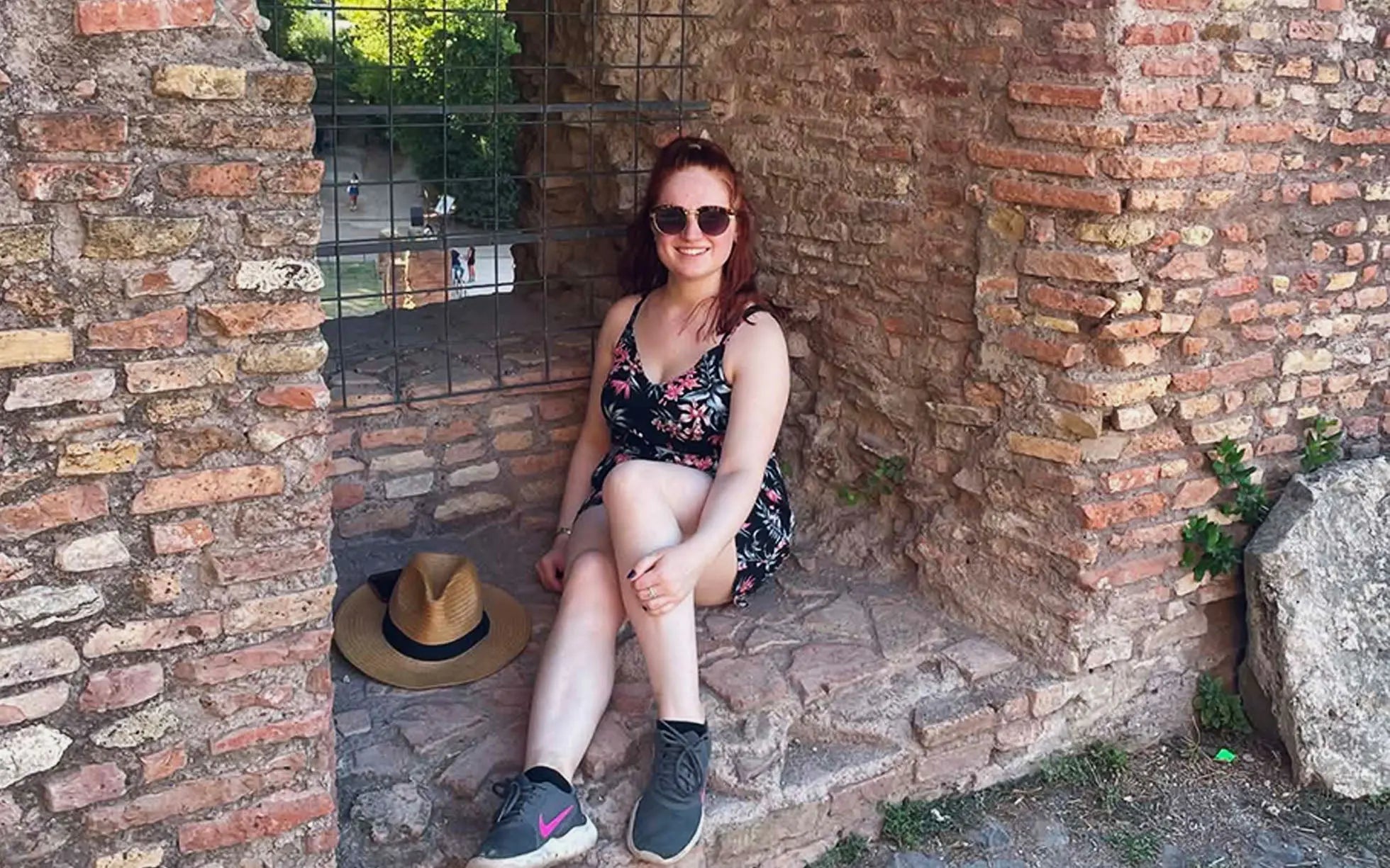 A woman sits in a stone archway surrounded by ancient brick walls, wearing sunglasses and a floral dress.
