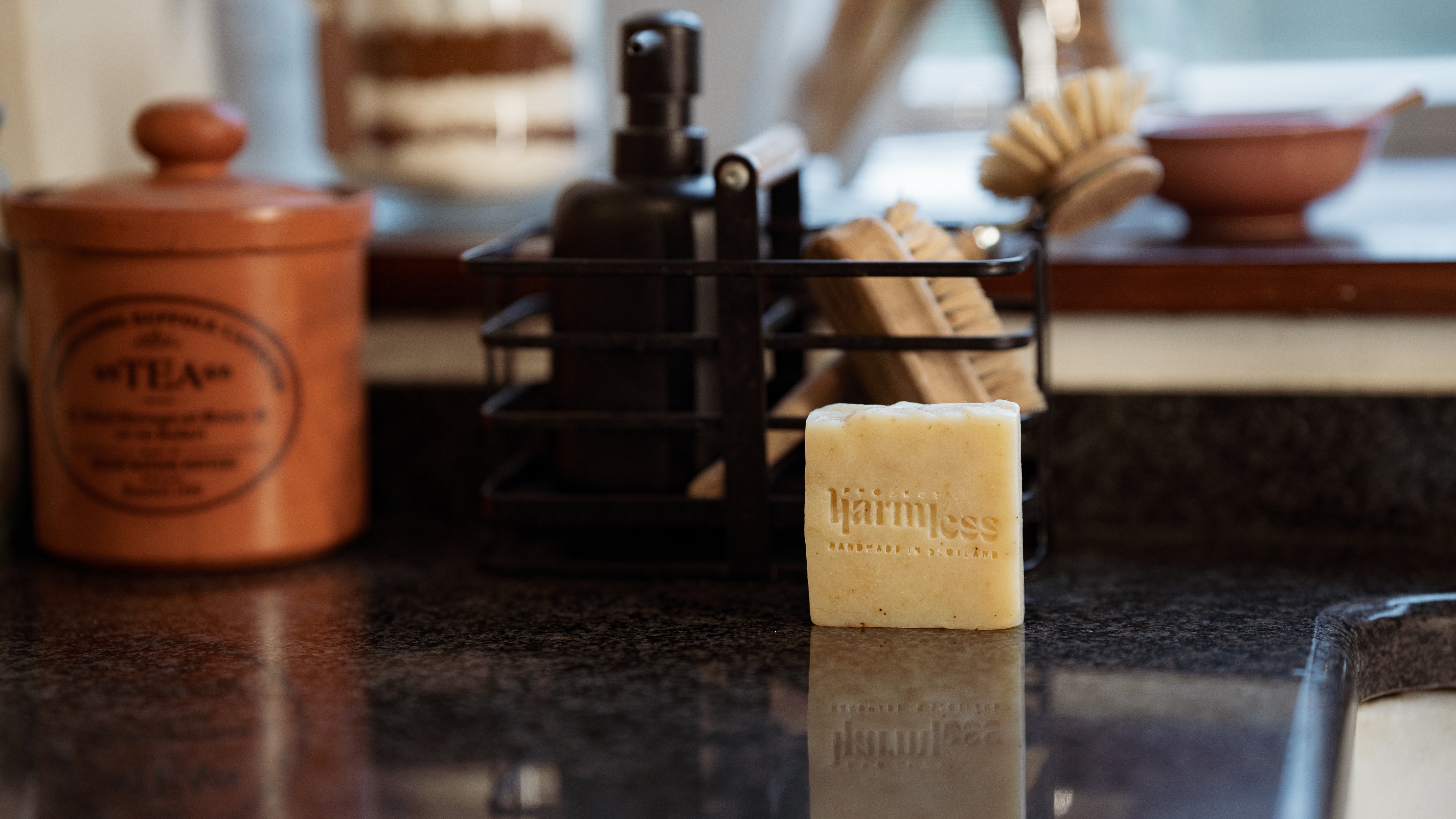 Project Harmless Scottish Lavender Soap resting on a beautifully curated sustainable kitchen countertop, surrounded by eco-friendly essentials. The soap is embossed with "Project Harmless" logo and features a "Handmade in Scotland" stamp, highlighting its artisanal craftsmanship. A refined, earth-conscious aesthetic with natural textures complements the soothing lavender theme.