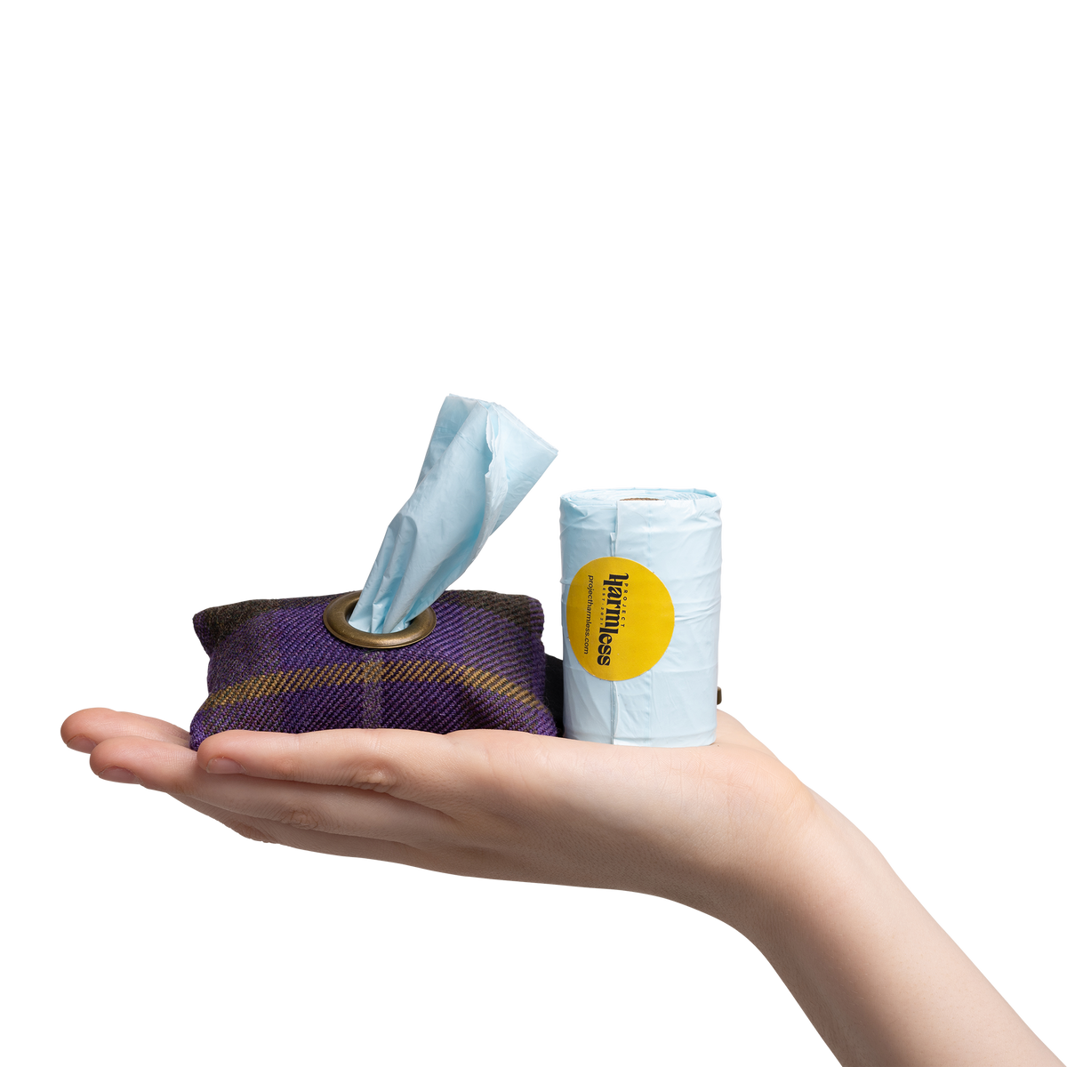 A hand holding a purple plaid limited edition purple tartan poo bag holder next to a roll of Harmless Poop Bag with a Project Harmless logo on it.