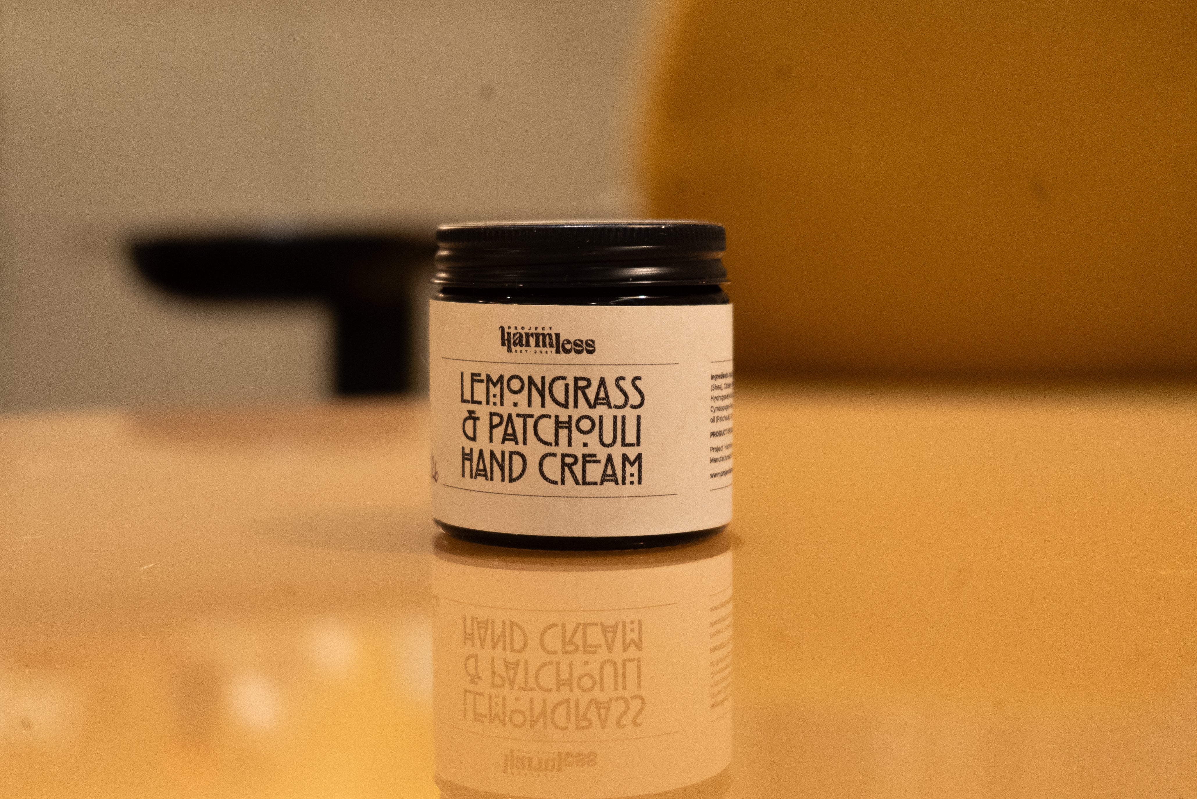 Jar of Project Harmless lemongrass and patchouli hand cream on a reflective surface with a blurred background